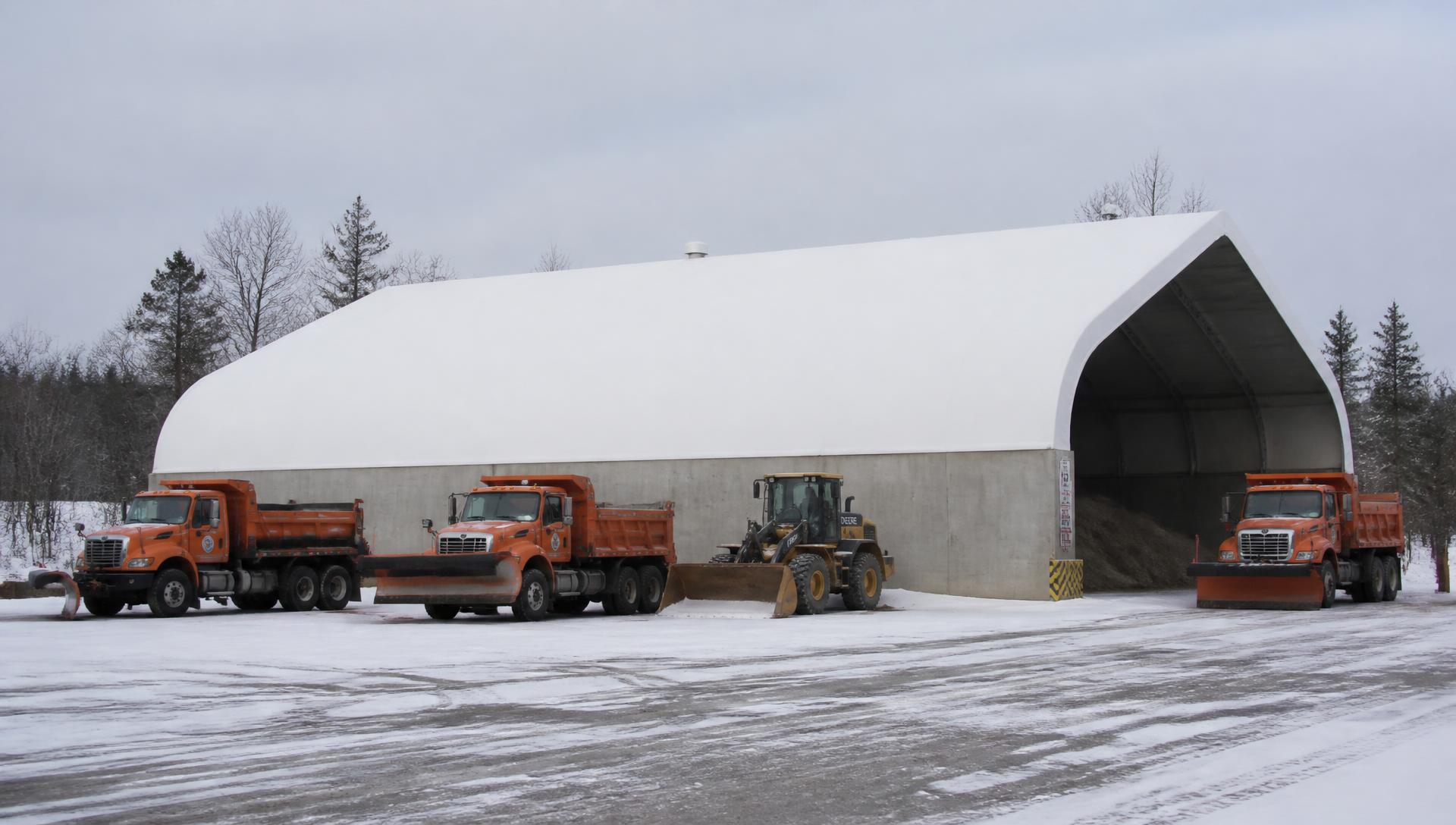 Orange municipal plow trucks and a loader outside a white fabric salt storage building on concrete stem walls in an Ontario public works yard in winter